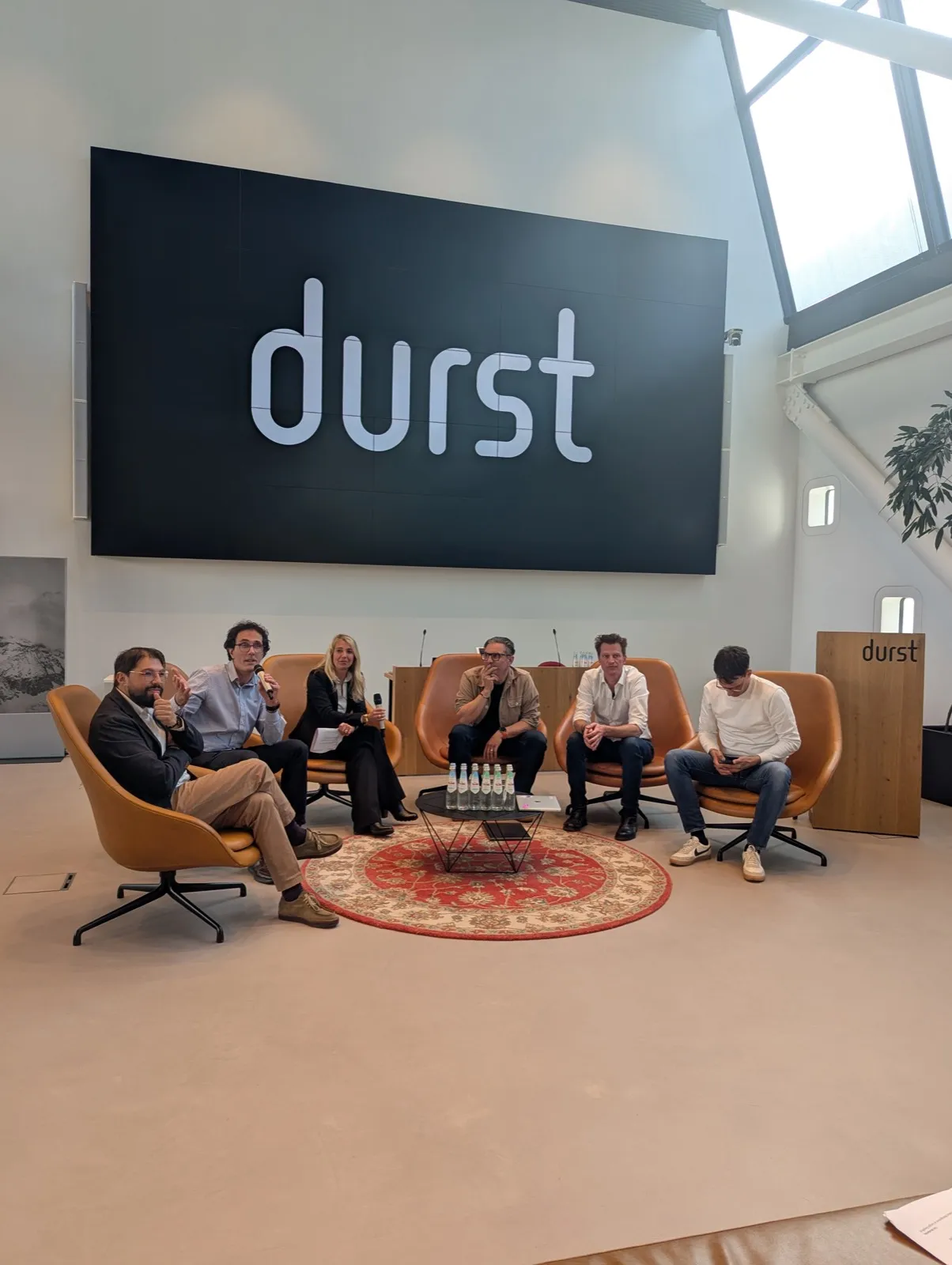 Six panellists seated on tan lounge chairs on a round patterned rug under a massive black wall sign reading durst inside a bright atrium