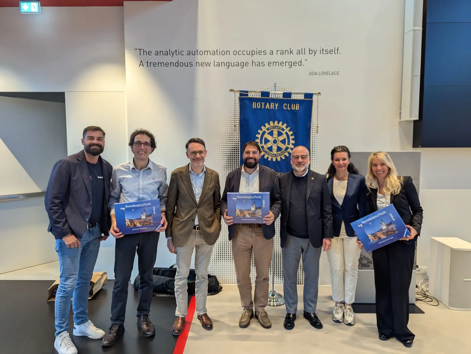 Seven people posing with printed calendars beneath a Rotary Club banner and a wall panel with a printed quotation about analytic automation