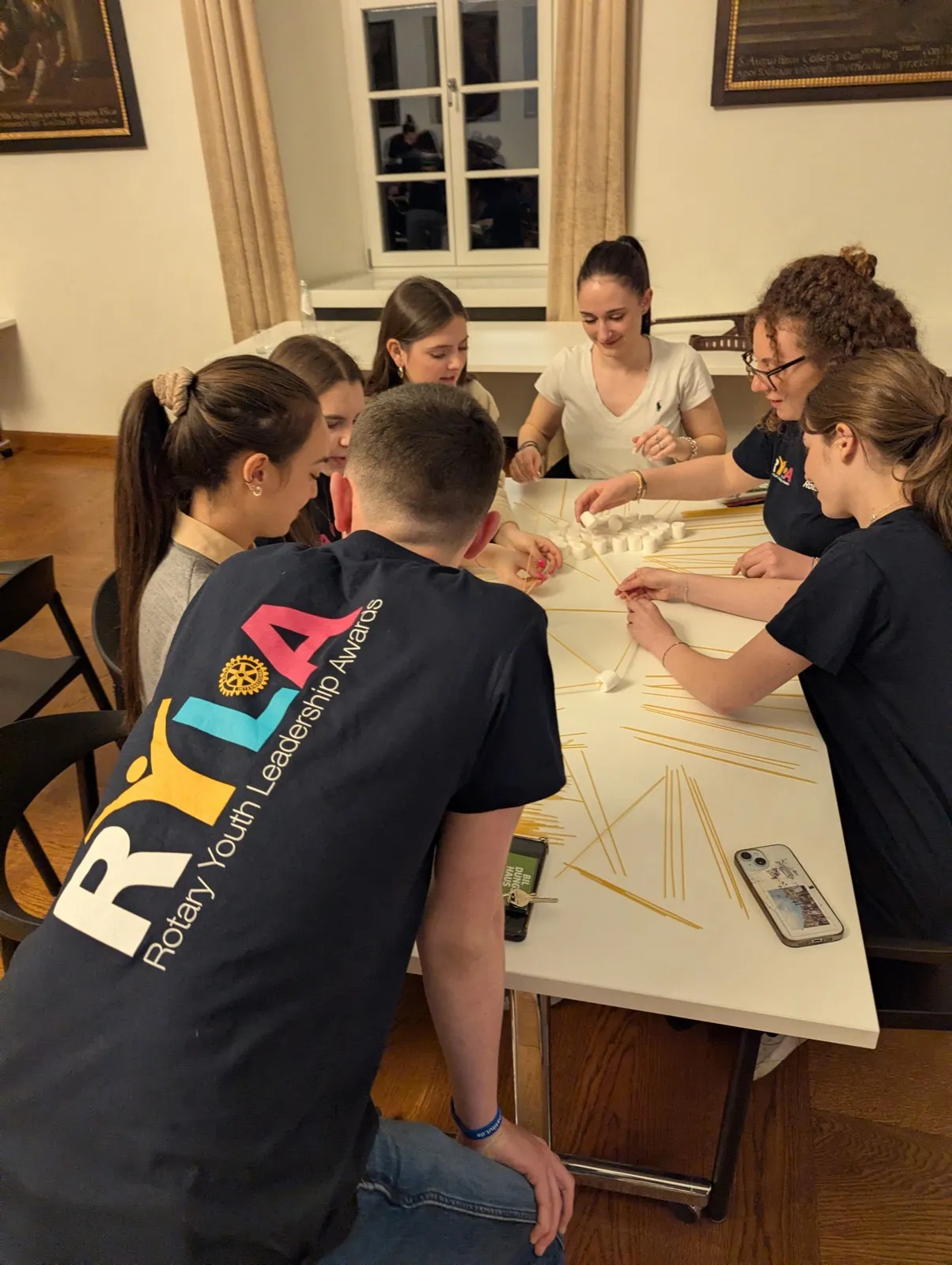 Seven teenagers wearing RYLA t-shirts building a tower from raw spaghetti sticks and marshmallows around a white table in a wood-panelled room