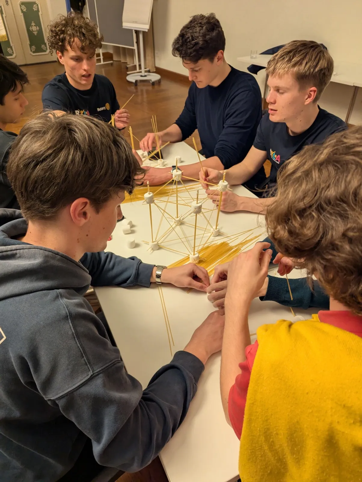 Six young men leaning over a table constructing an intricate spaghetti and marshmallow lattice, focused on connecting joints with careful hands