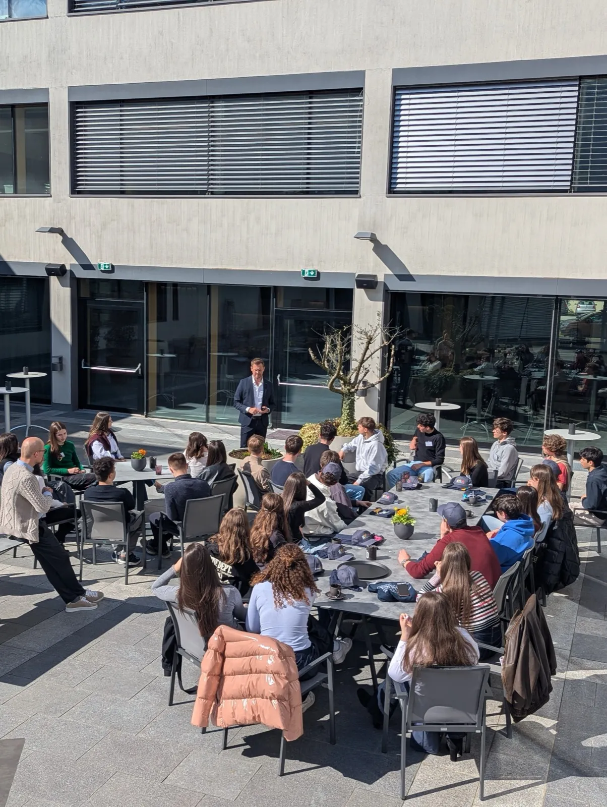Participants seated together at outdoor terrace tables in the sun, talking beside a low glass balustrade