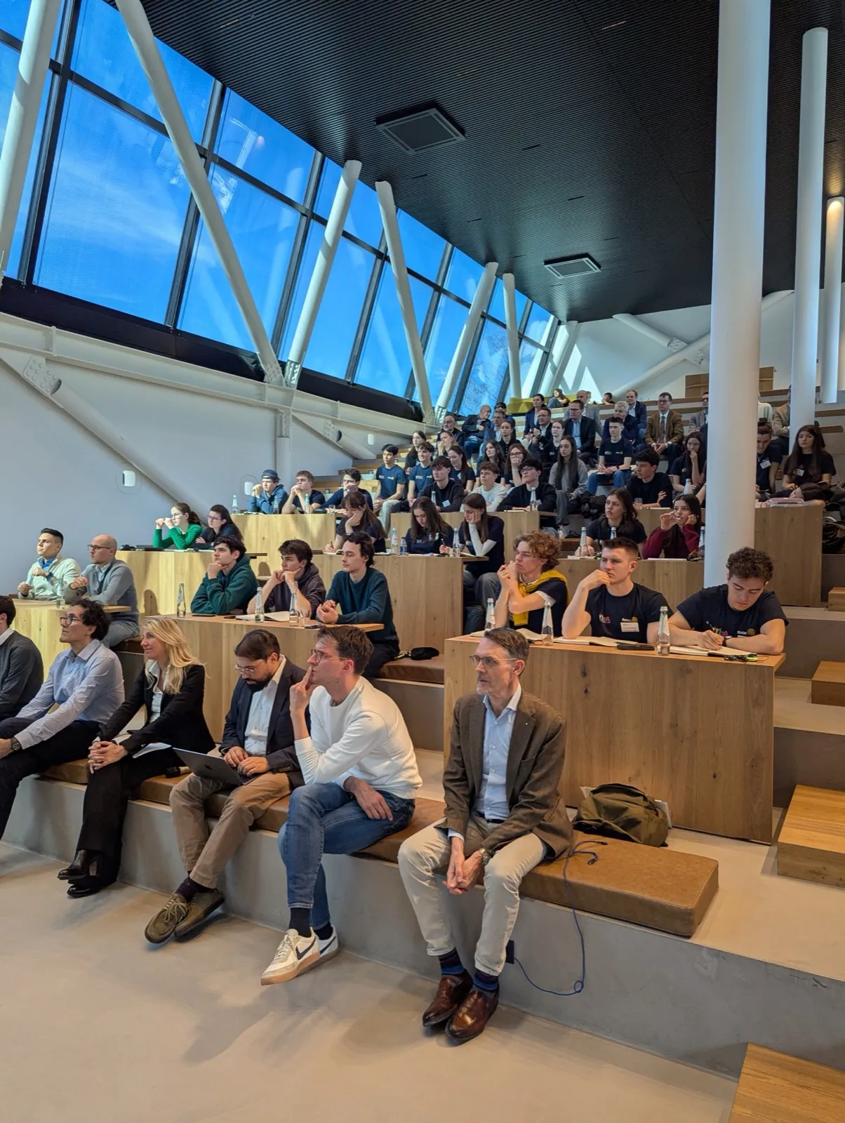 A wide auditorium beneath a slanted glass roof, filled with participants seated on tiered wooden steps