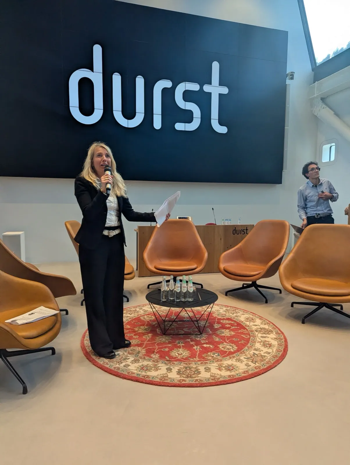 A speaker with a microphone gesturing on a stage beside a lectern, in front of rows of empty teal chairs