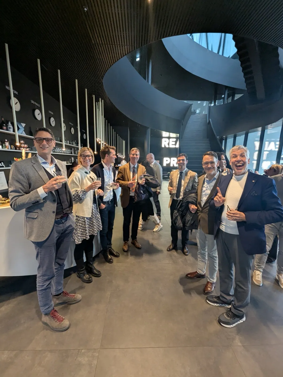 Organisers and guests in jackets raising glasses together in a dark modern atrium beneath a sweeping spiral staircase