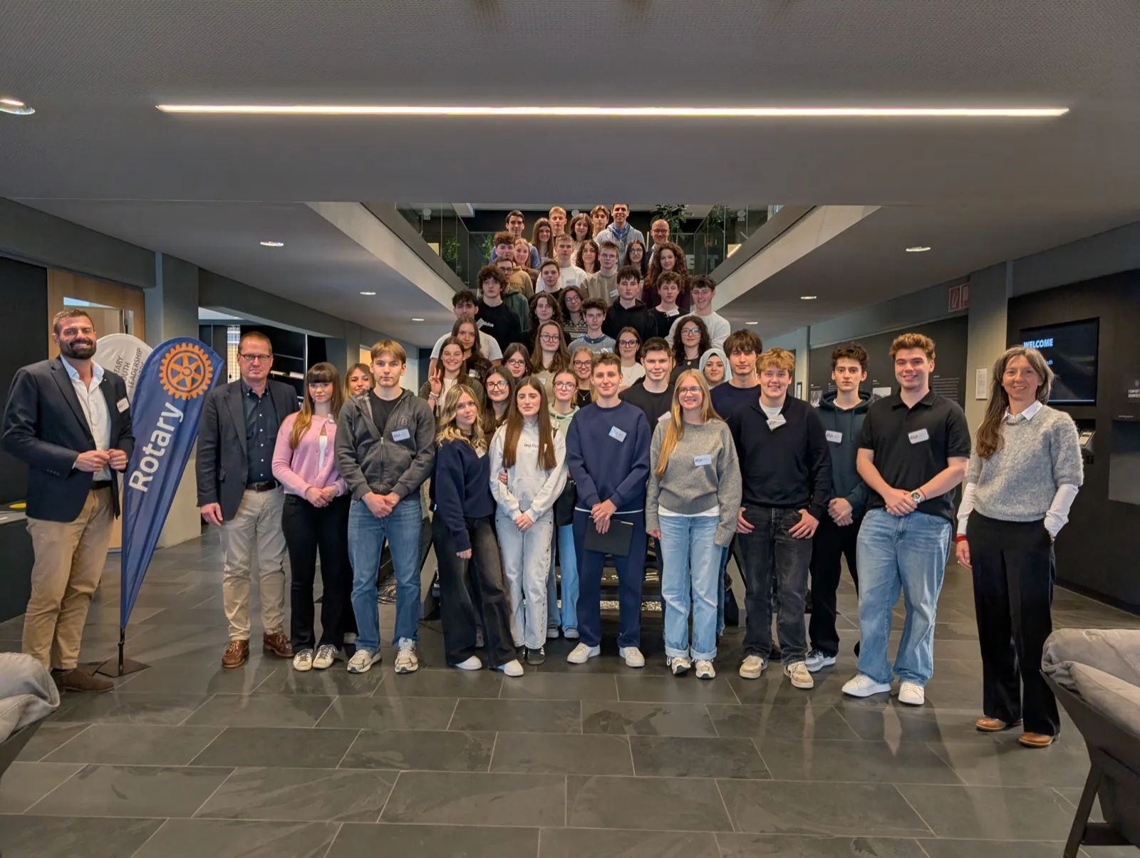 Large group portrait of teenagers standing together beside a blue Rotary banner inside a modern lobby with polished slate flooring