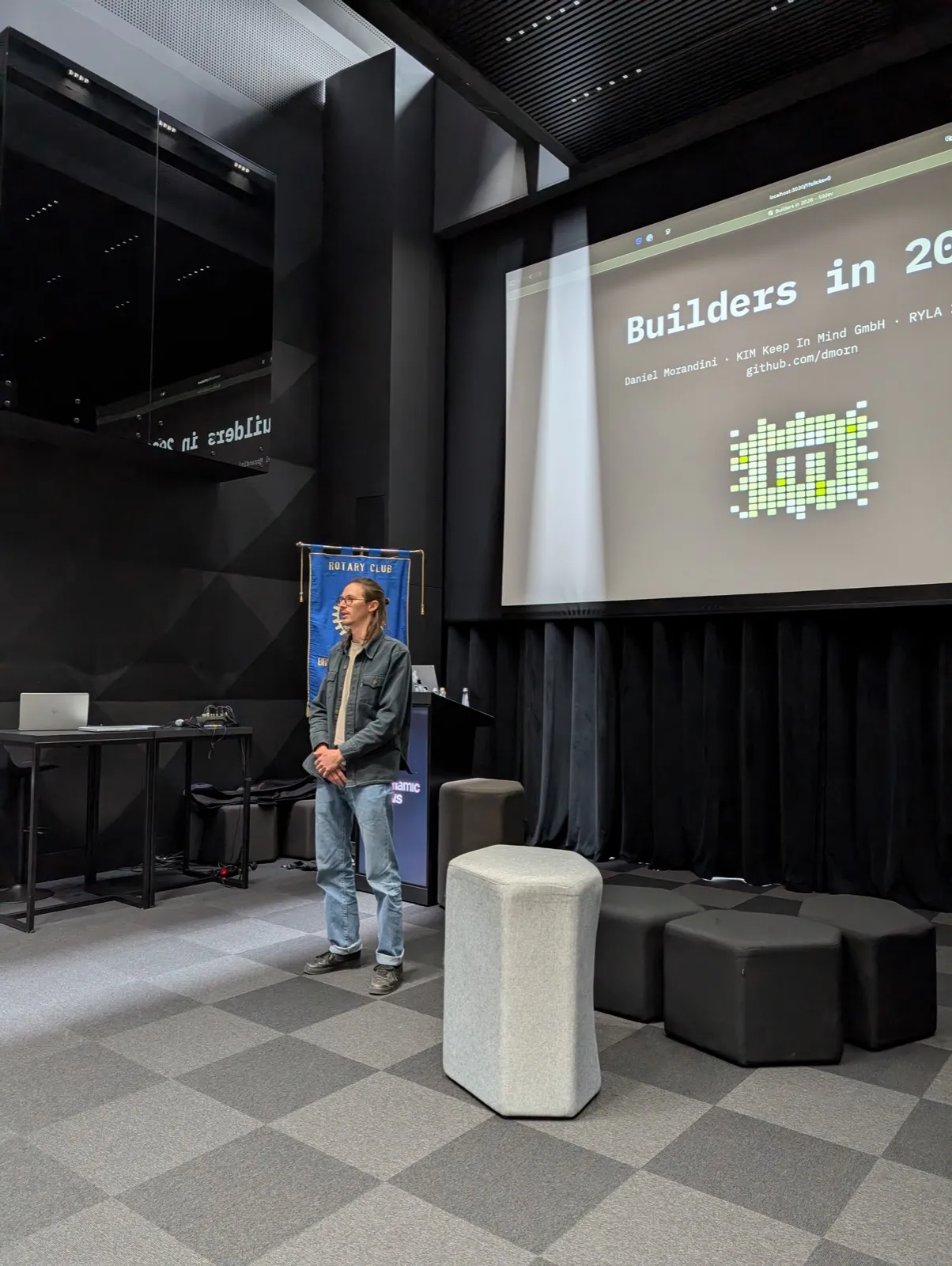 A bearded speaker in a denim jacket standing beside a lectern with a projected slide reading Builders behind him in a dark auditorium