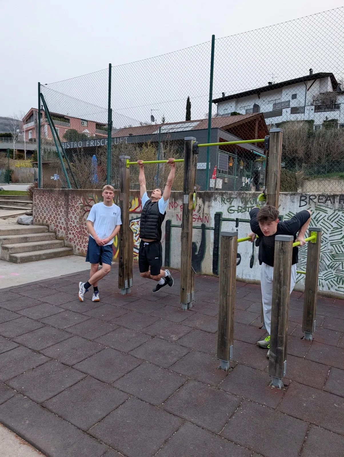 Three young men training at an outdoor calisthenics park with wooden bars and graffiti walls, houses and mountains rising in the background