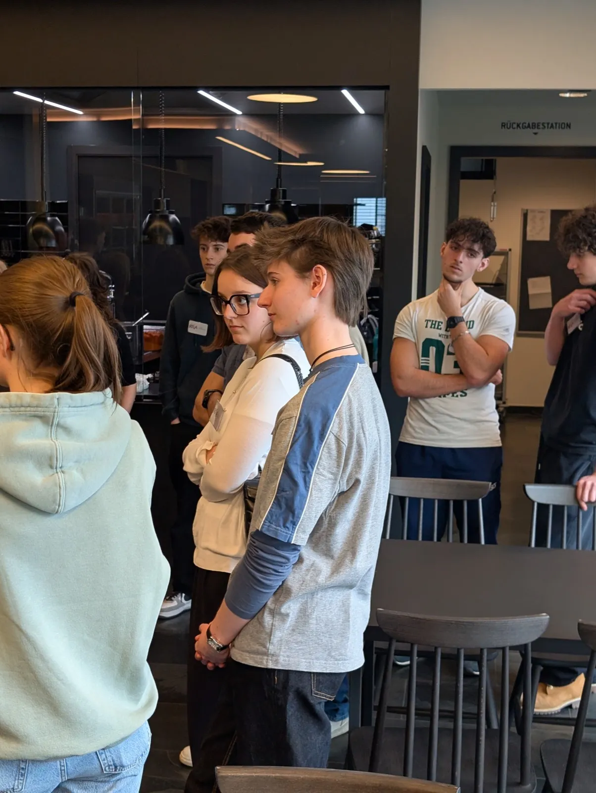 Youths standing and listening attentively near a glass-walled cafeteria area with a small return-station sign visible on the wall