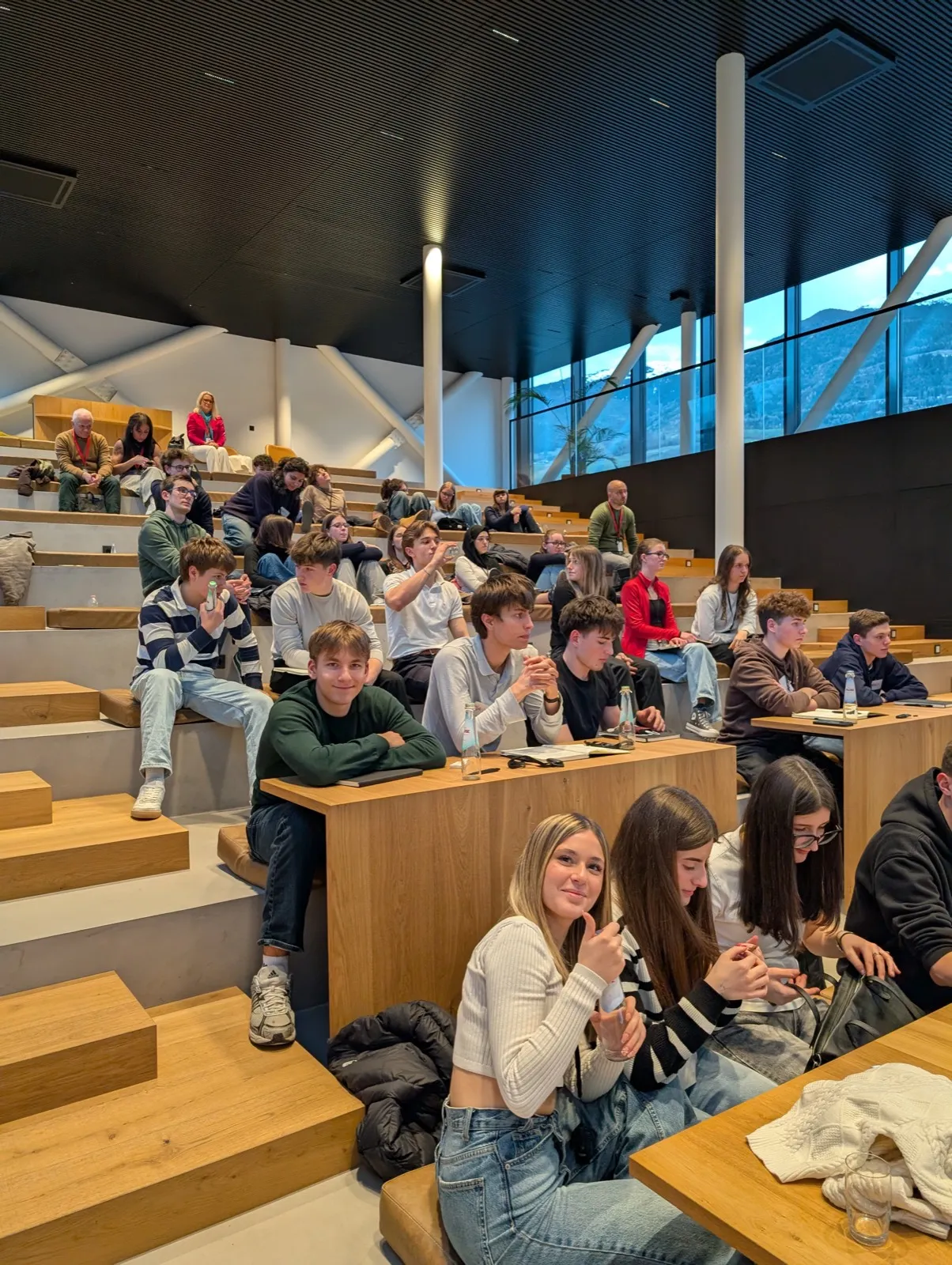 Teenagers seated on tiered wooden benches inside a bright lecture hall with panoramic windows revealing mountains under a grey sky outside