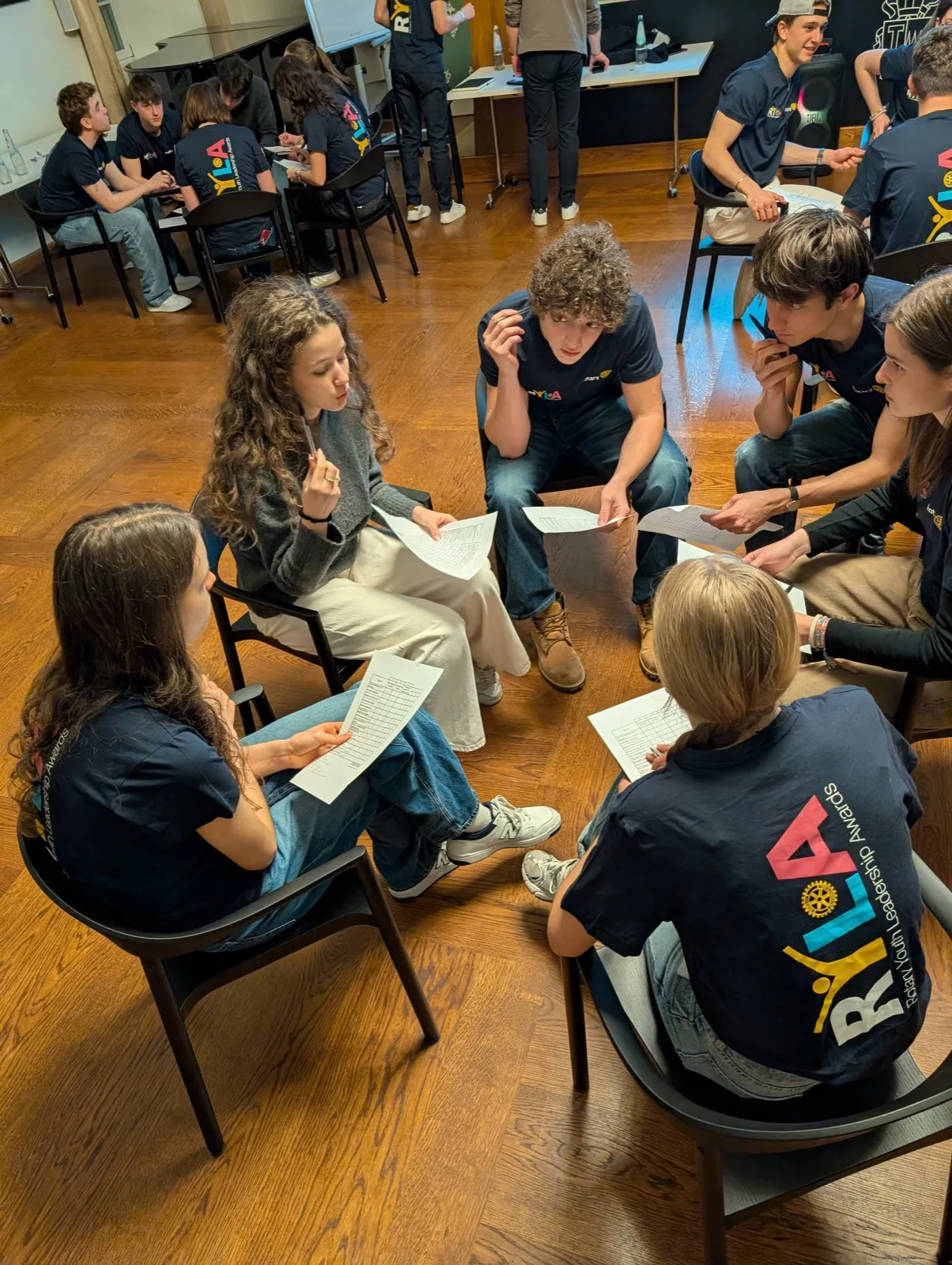 Overhead view of young participants in dark shirts sitting in a circle on chairs, reading printed sheets during a group workshop discussion