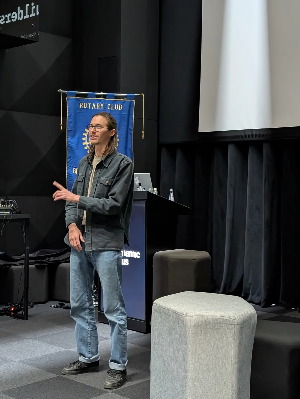 A long-haired speaker in a denim shirt gesturing beside a blue banner and lectern on a dark stage with grey carpeting and cushioned seats
