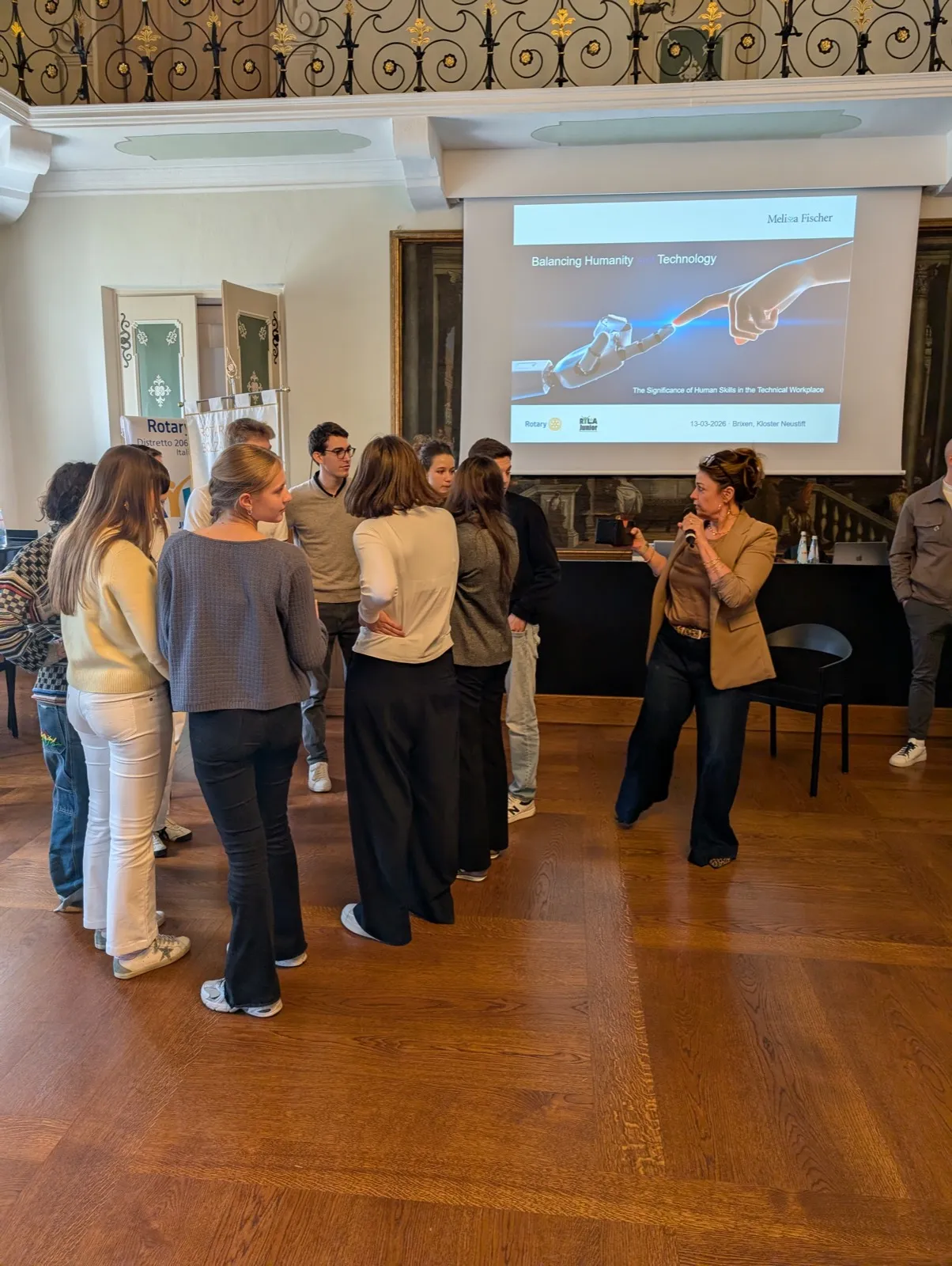 A woman with a microphone addressing a semicircle of young listeners in a stately hall, a projected slide reading Balancing Humanity Technology behind her