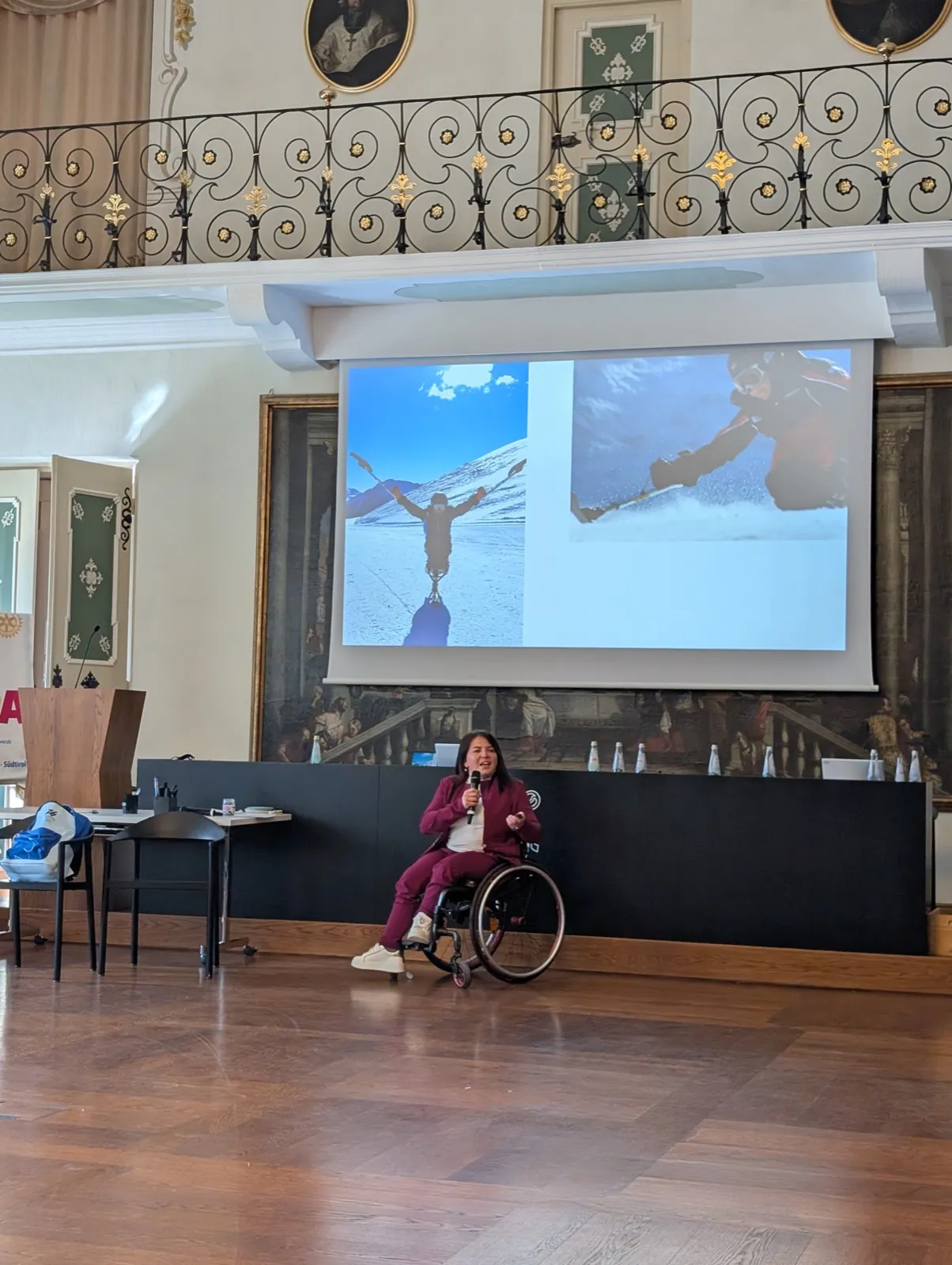 A woman in a maroon tracksuit sitting in a wheelchair holding a microphone below a projected slide showing snowy mountain skiing scenes on the wall