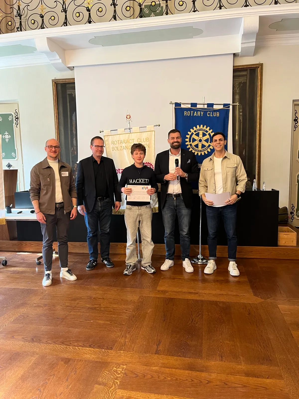 A group of young men lined up holding certificates beside a Rotary banner and classical portrait paintings inside an elegant wood-floored reception room