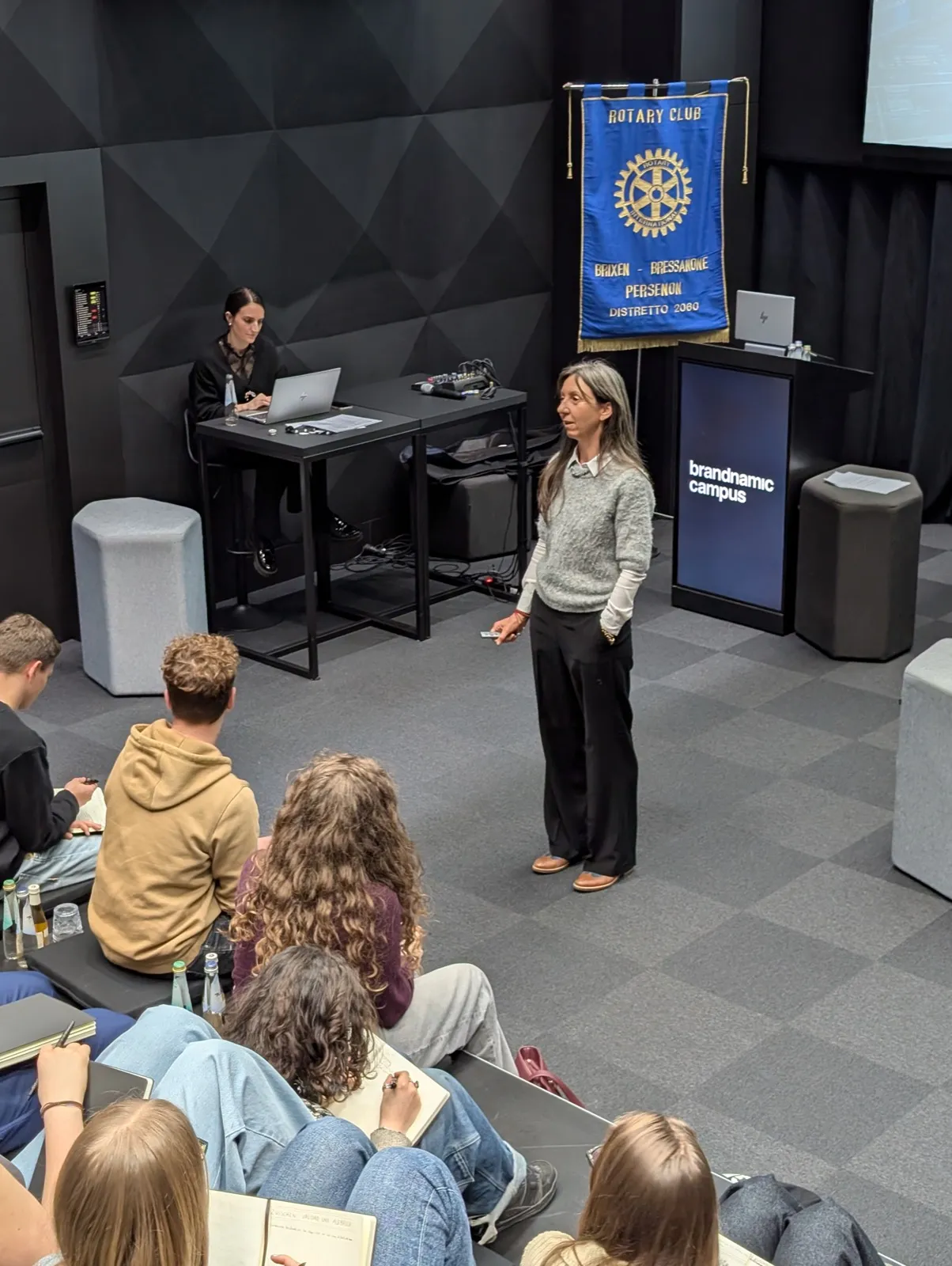 A woman in a grey knit sweater speaking to a seated audience beside a hanging Rotary banner and dark lectern displaying the text brandnamic campus