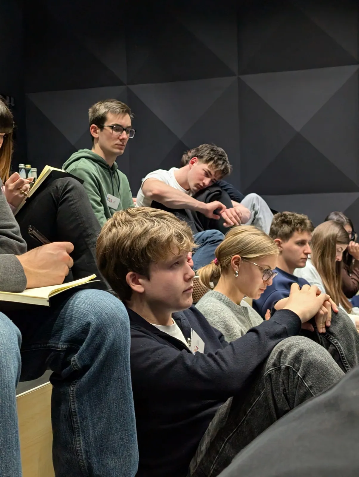 Youths seated closely together taking notes attentively in a dimly lit acoustic-panelled room, one wearing glasses and a green hoodie in focus