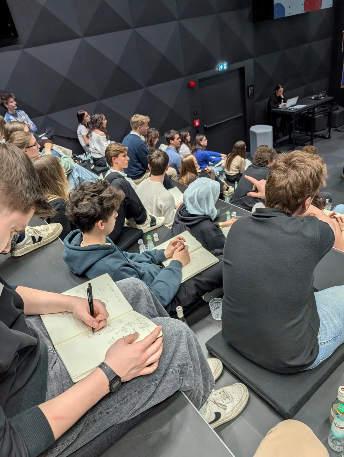 Rows of young participants writing in notebooks on the tiered steps of a modern auditorium while a facilitator works at a distant desk