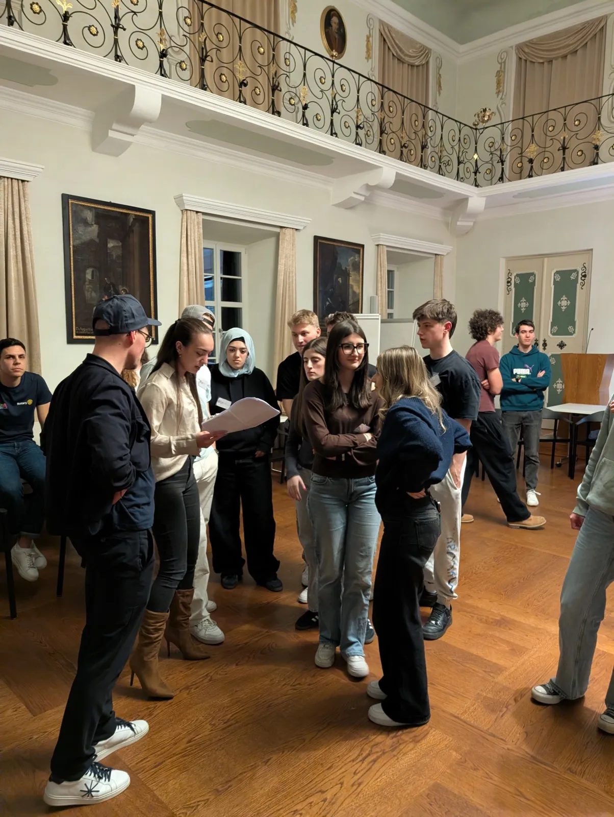 A small circle of standing teenagers looking at a paper held by a young woman under a wrought-iron gallery railing and historic paintings