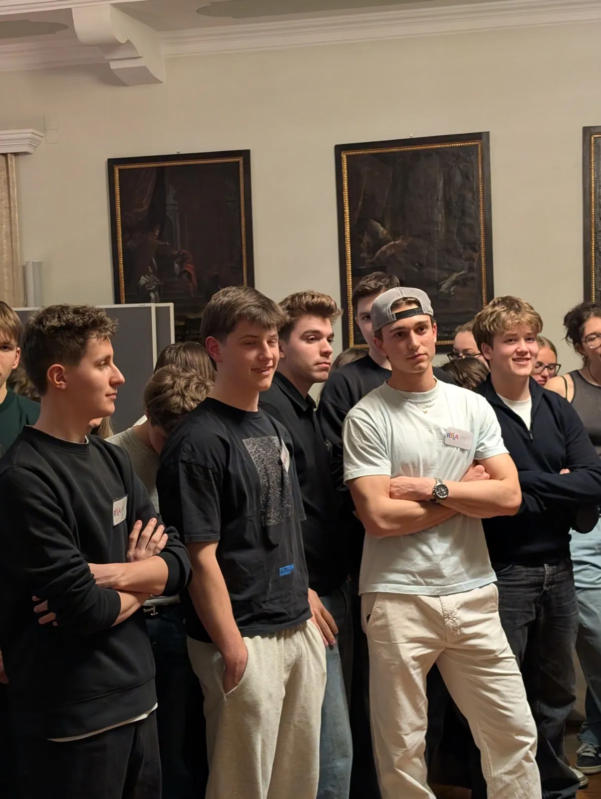 A group of young men clustered together with crossed arms standing before large gilt-framed dark oil paintings inside a warmly lit historic room