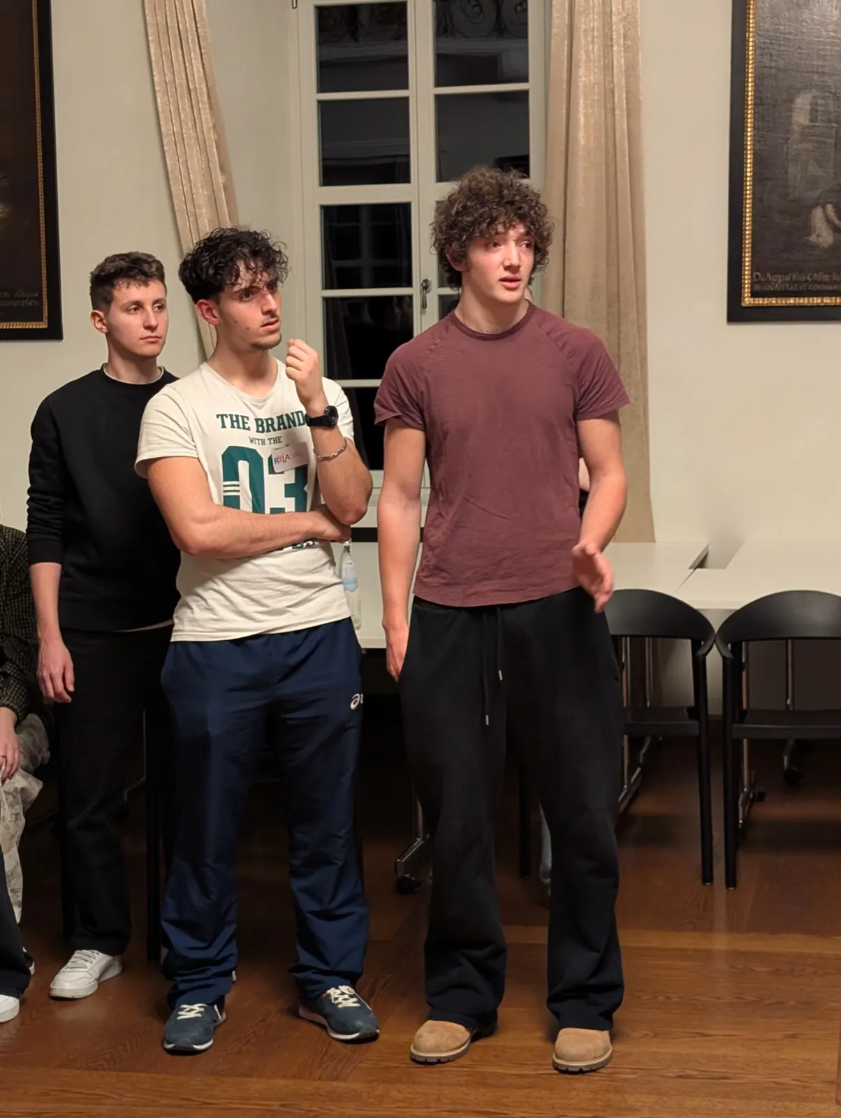 Two curly-haired young men speaking in a candlelit room with framed old documents on the wall behind them and empty stacked chairs