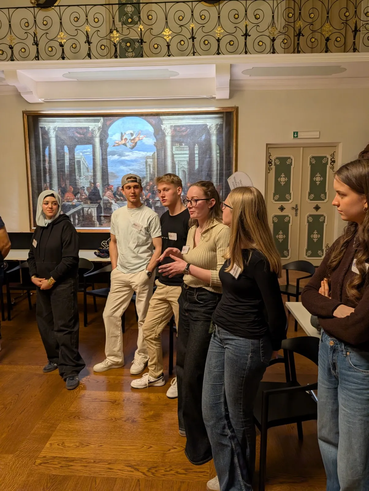A cluster of young participants conversing beside a large classical fresco of columns and cherubs mounted above a bench in a heritage hall