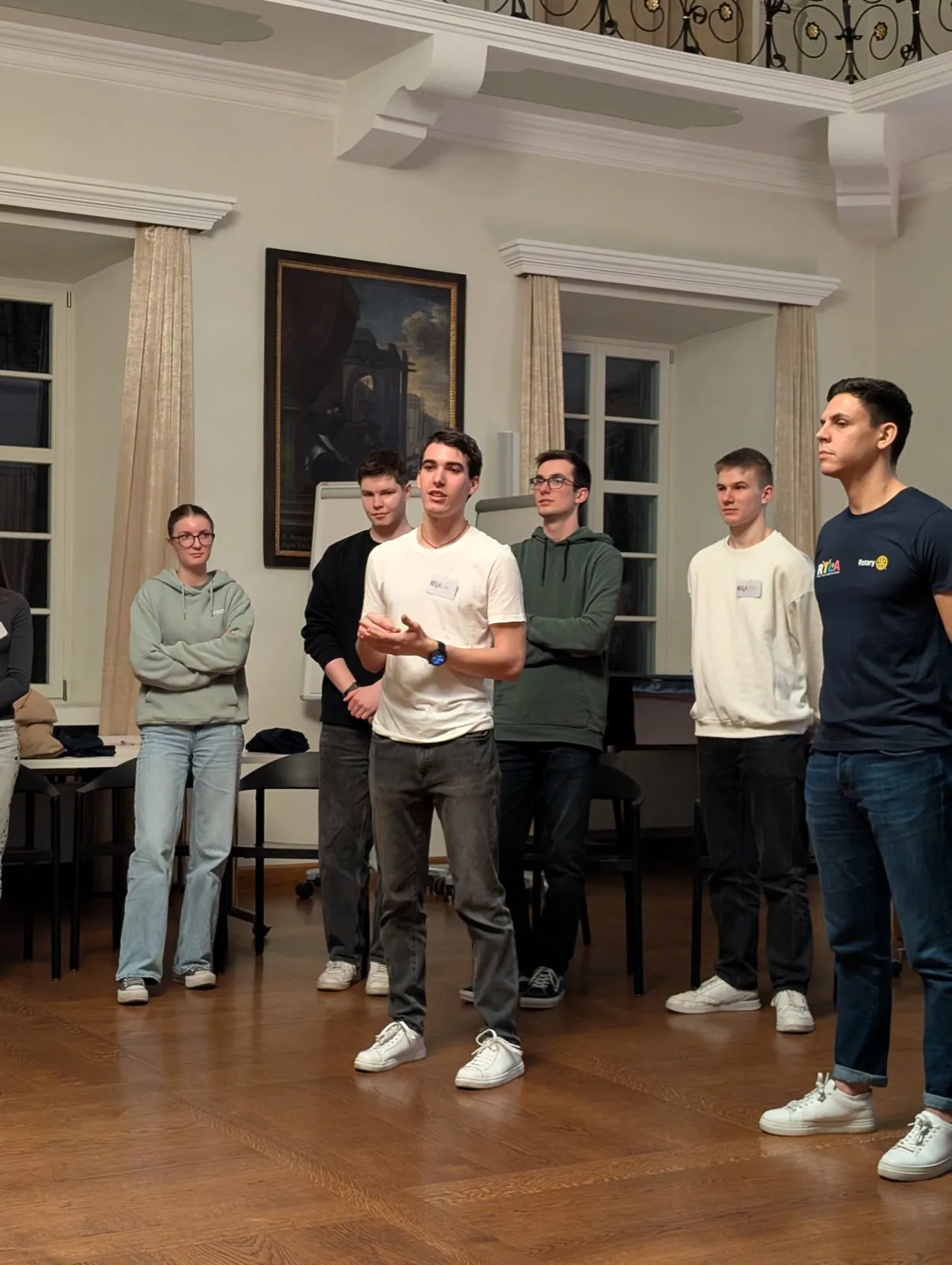 A line of young men standing on a wooden parquet floor speaking to unseen listeners with a dark oil painting hung on the wall behind them