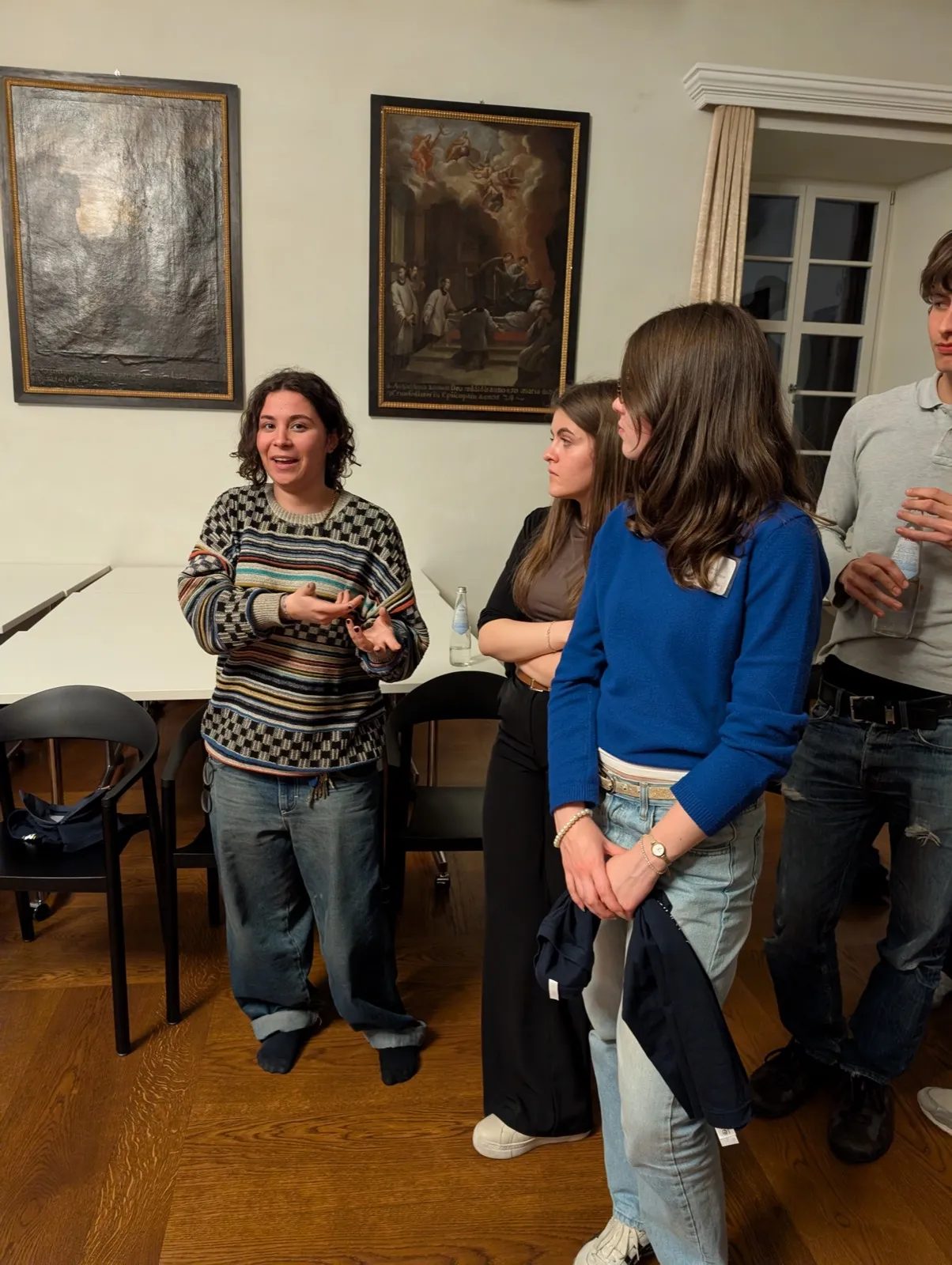 A young woman in a patterned sweater gesturing while talking to two peers, with religious baroque paintings hanging on the wall behind them