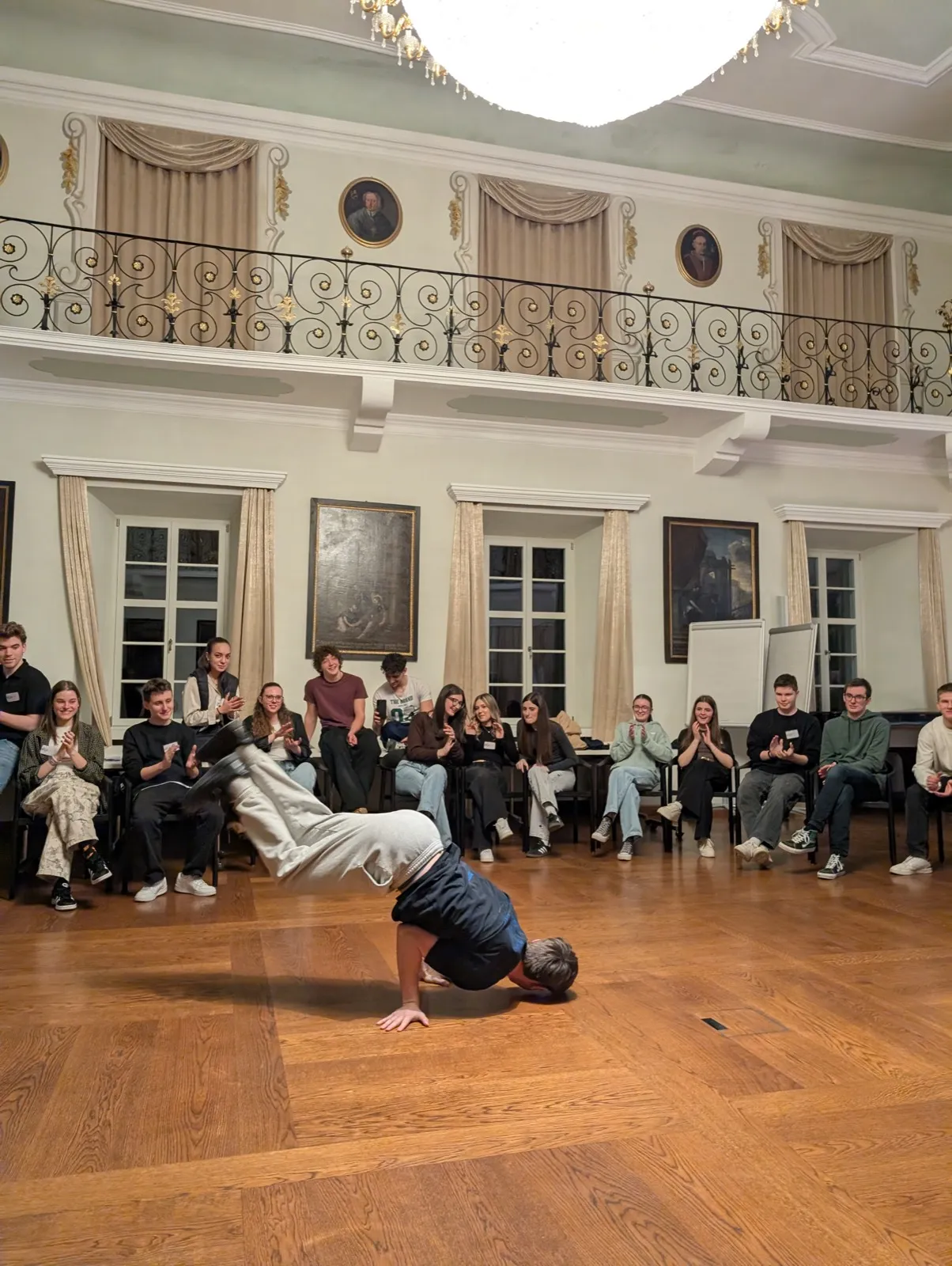 A break-dancer balancing on one shoulder mid-move on parquet floor while seated peers clap along the perimeter of a chandelier-lit hall