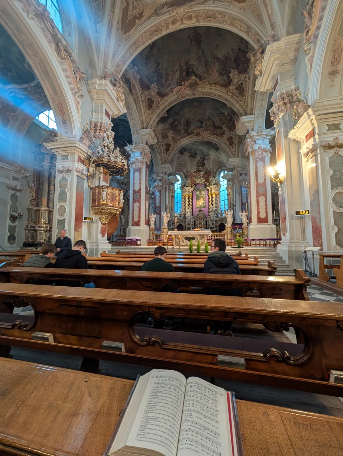 A baroque church seen from the back with vaulted frescoes, a gilded altar, visitors seated in dark wooden pews, and an open songbook resting nearby