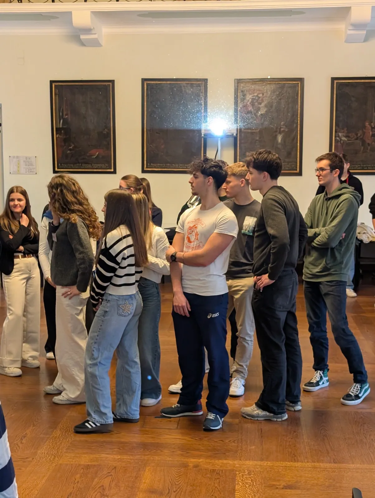 A cluster of teenagers standing loosely and chatting on parquet flooring beneath a row of dark gilt-framed historic paintings inside a bright hall