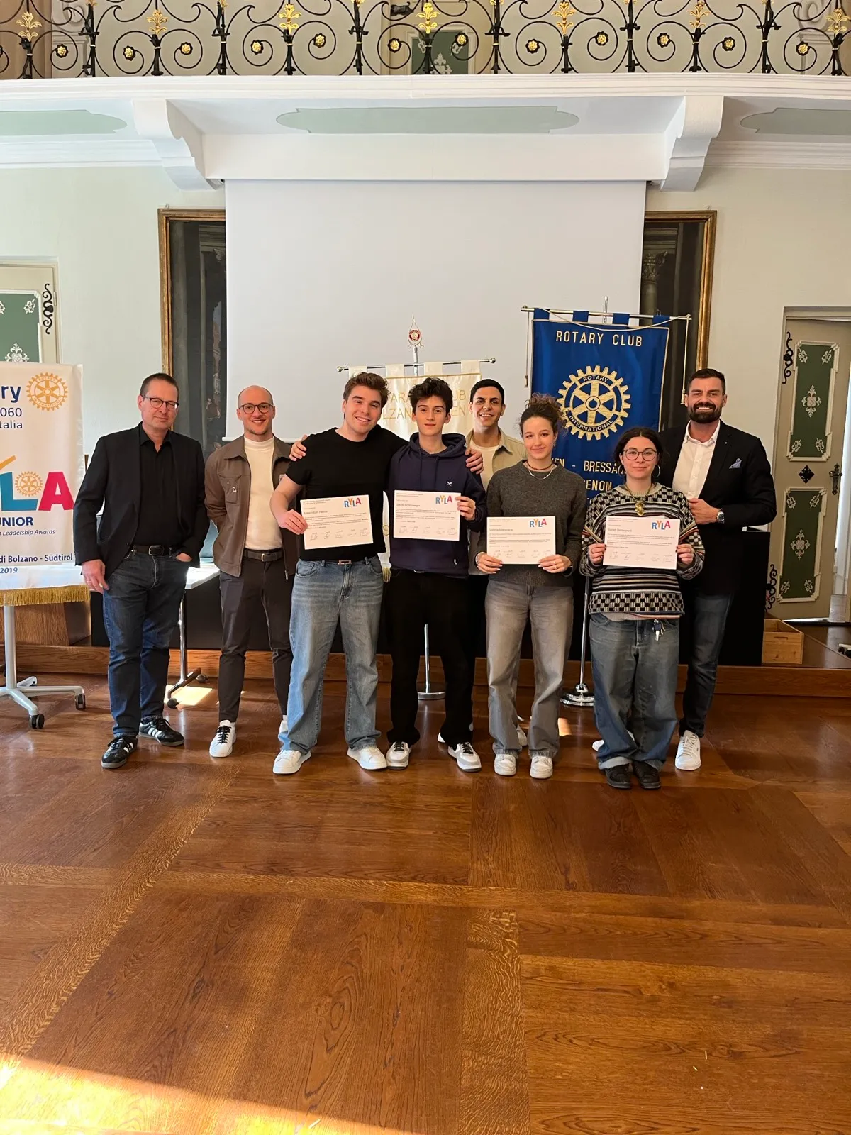 A row of young men holding printed certificates standing beside a blue Rotary banner in an elegant room with polished floorboards