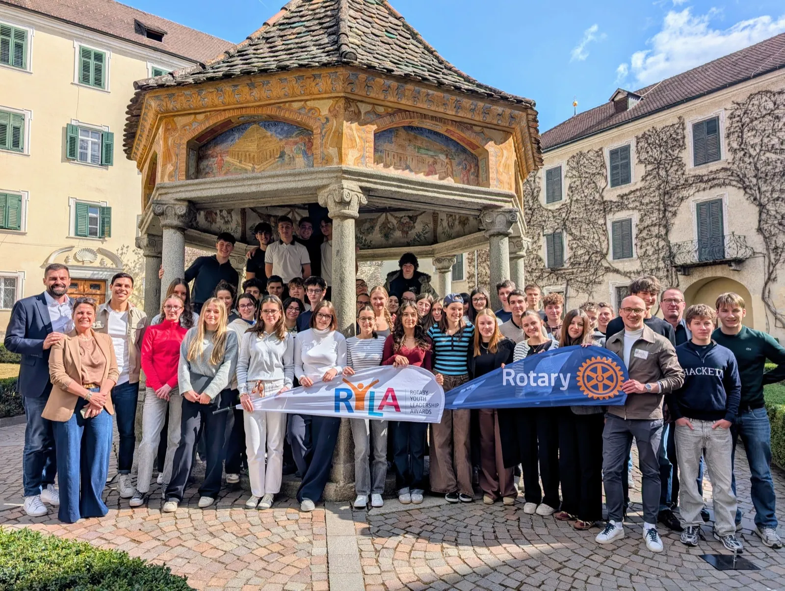A large group posing outdoors in front of the frescoed well pavilion of the monastery courtyard, holding the RYLA and Rotary banners, under a blue sky