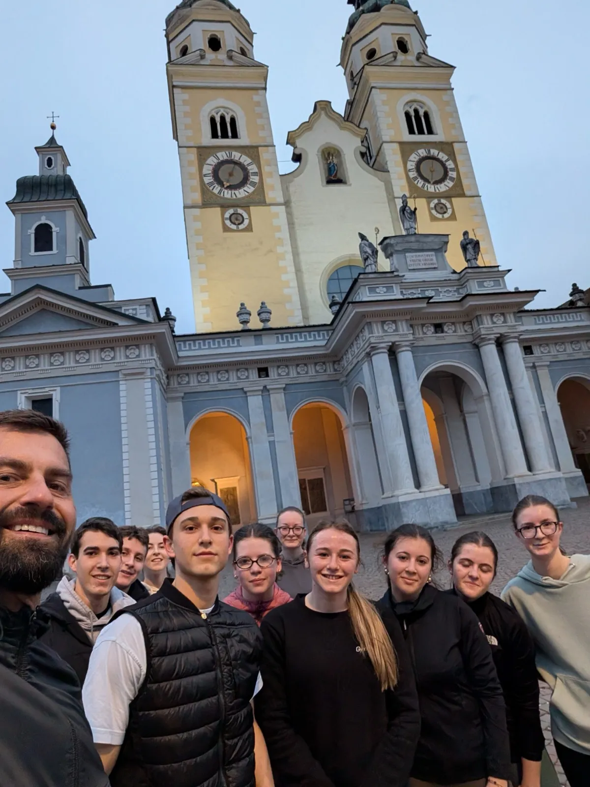 A group selfie of participants and an organiser in front of a floodlit church with twin clock towers at dusk