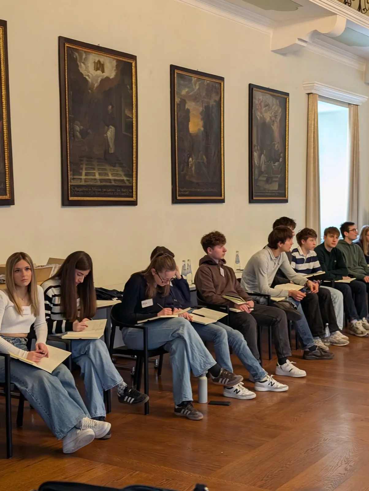 A row of participants seated with open notebooks beneath dark framed paintings, listening in a bright hall