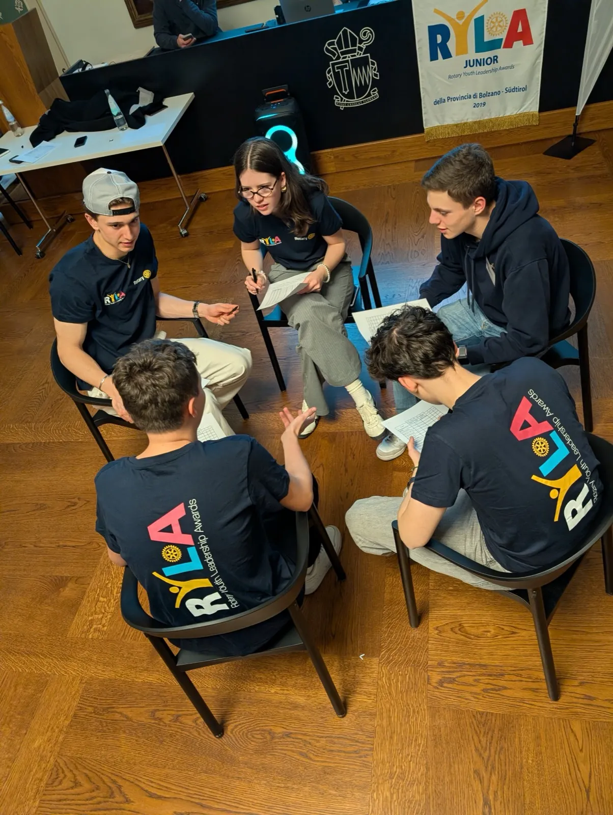 An overhead view of participants in matching RYLA shirts seated in a circle on a wooden floor during a discussion