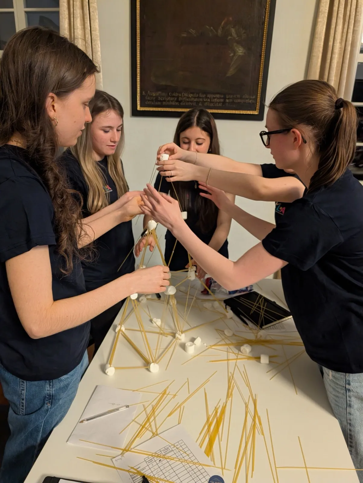 A group of participants reaching in together to place a marshmallow atop a tall spaghetti tower beneath a framed painting