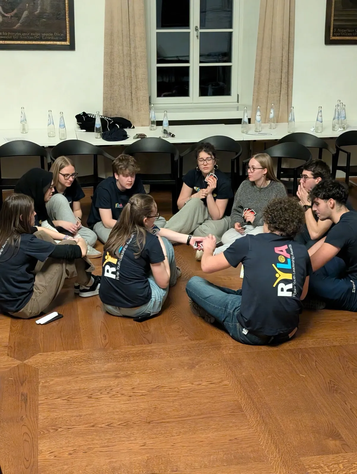Participants sitting in a circle on the floor playing a card game together beside a row of water bottles in the evening
