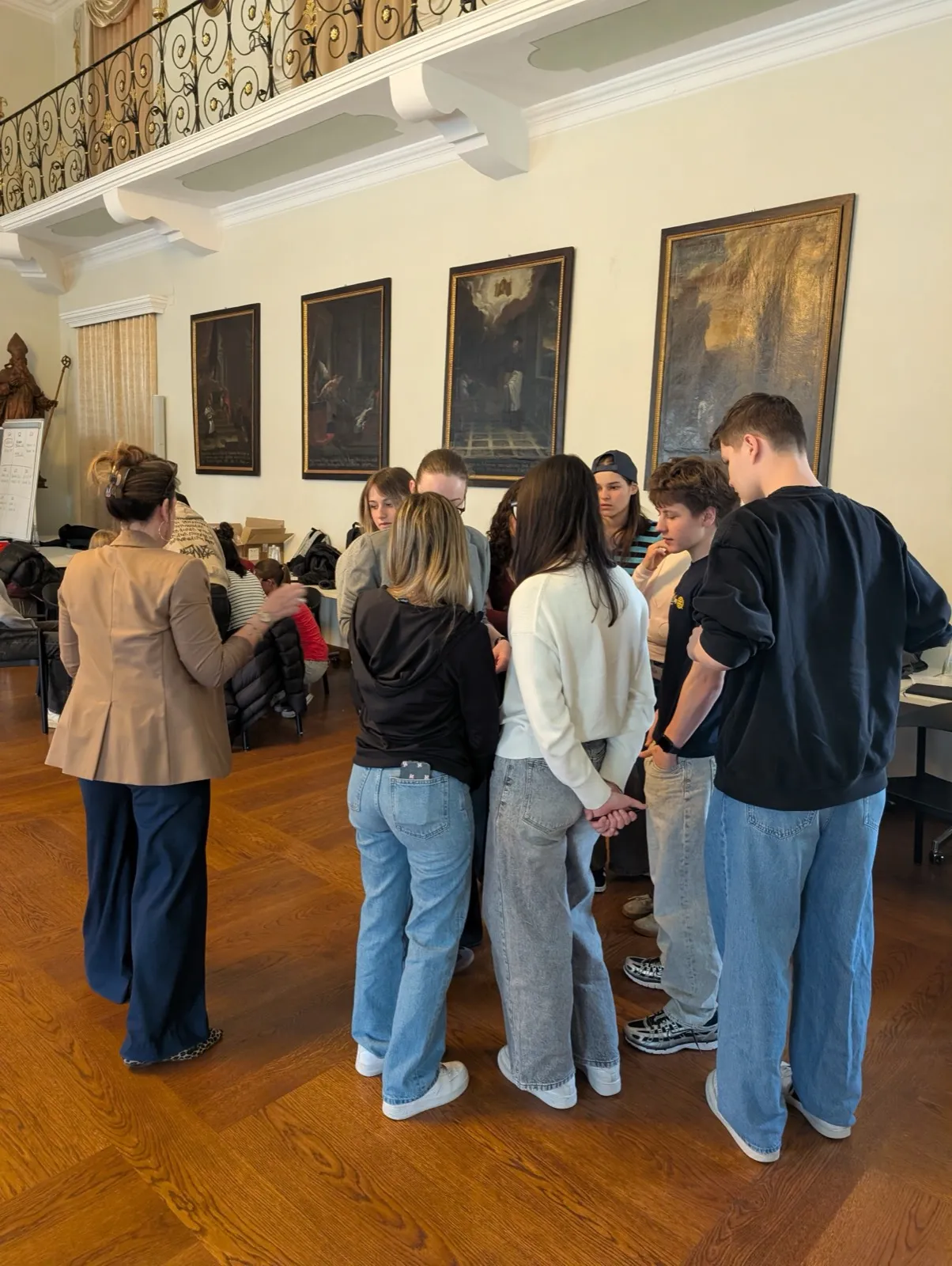 A small huddle of participants gathered close together in conversation beneath historic paintings, an organiser looking on