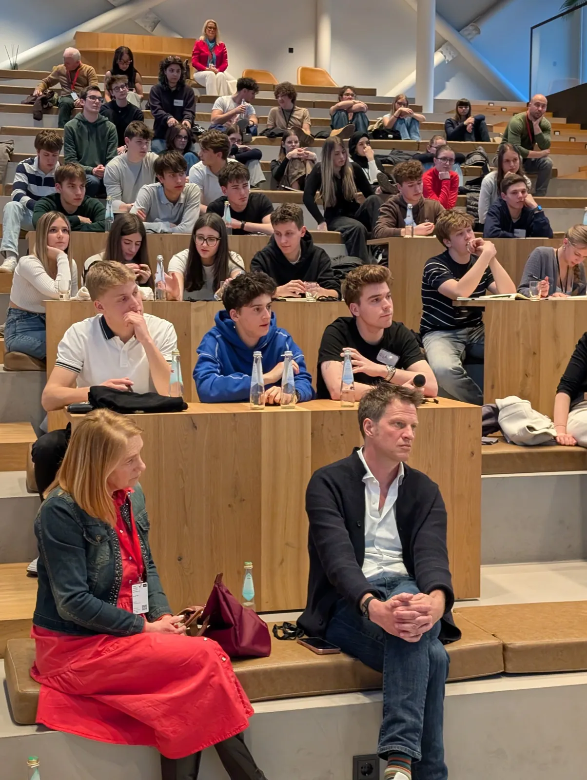 A tiered wooden auditorium filled with participants and adults, water bottles on the desks in front of them