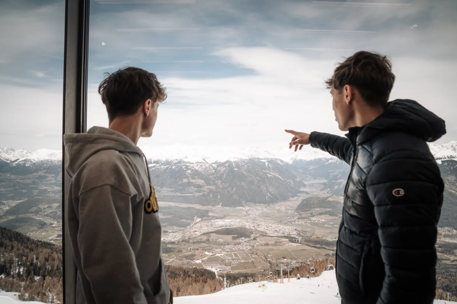 Two participants at the window of a cable car, one of them pointing out over a valley with snow-covered mountains behind it