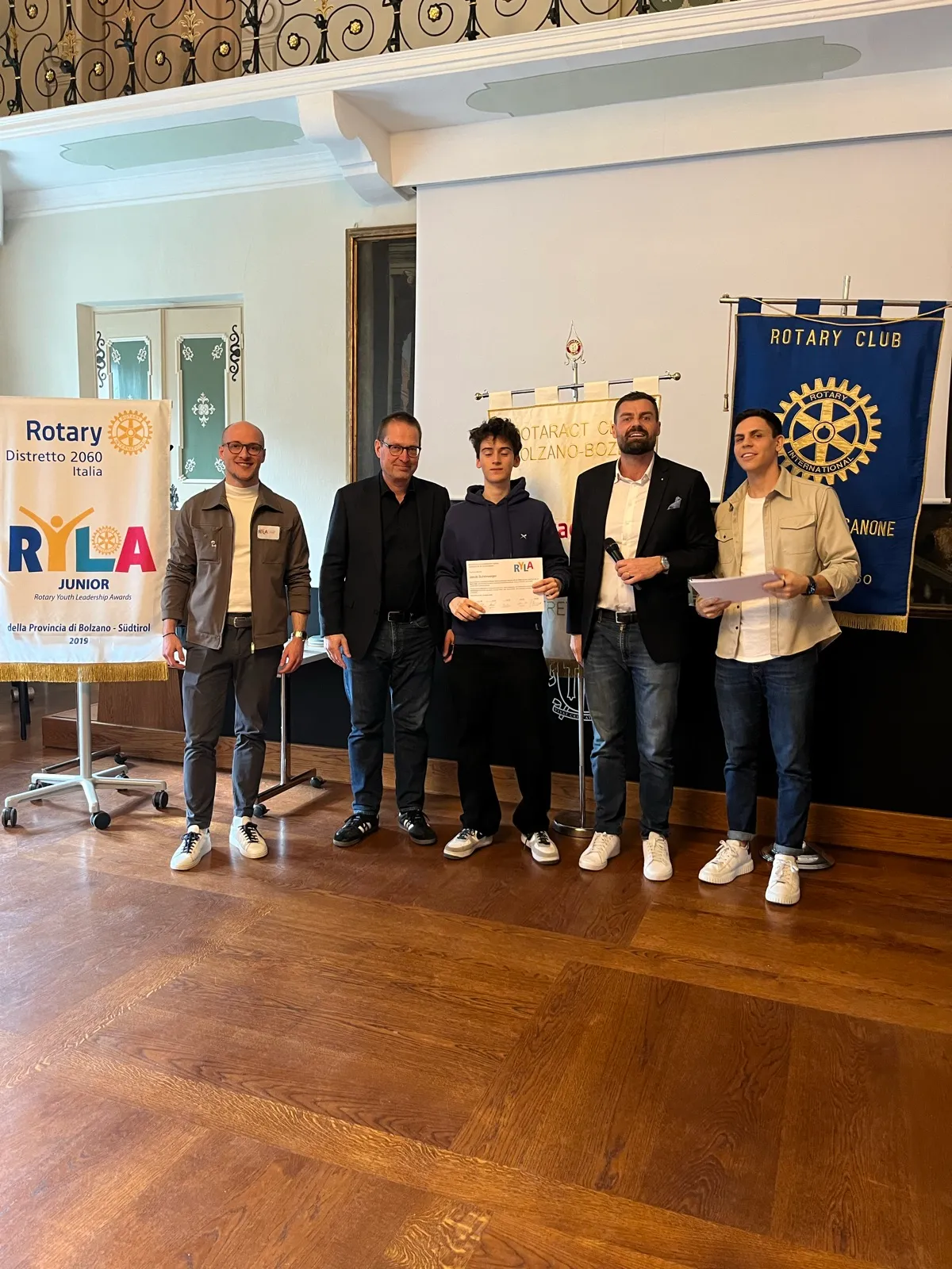 A participant holding a certificate between four organisers, in front of a RYLA Junior banner of Rotary District 2060 and a Rotary Club banner