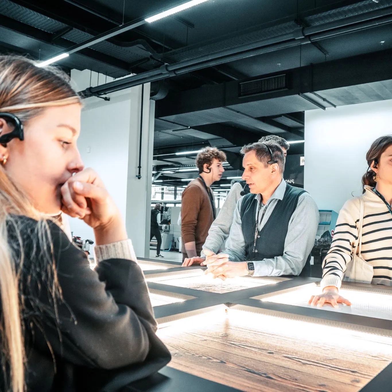 Students wearing tour headsets listening to a guide at an illuminated inspection table during a company visit