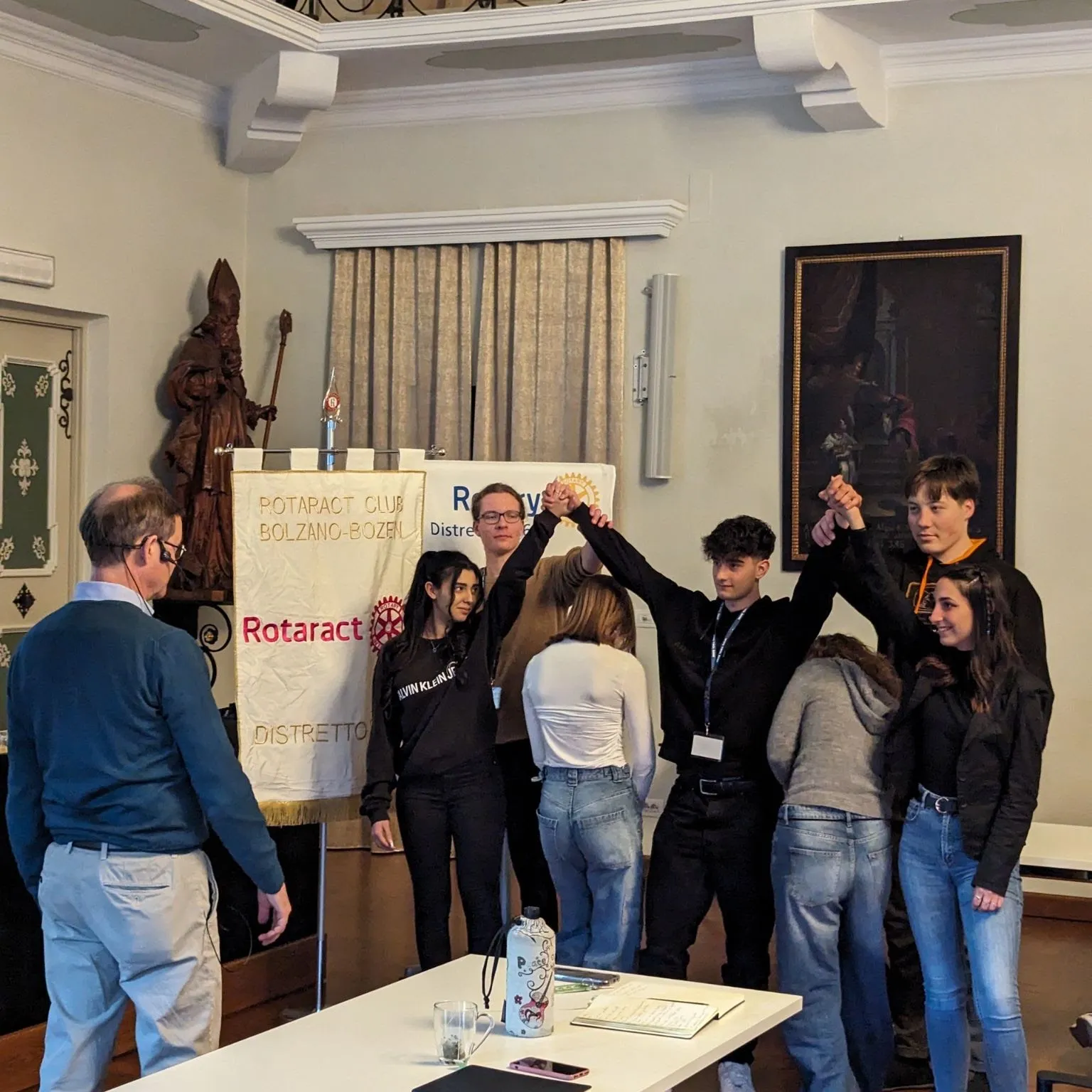 Students standing in a row with their raised hands joined into an arch, in front of a Rotaract Club Bolzano-Bozen banner, while an organiser looks on