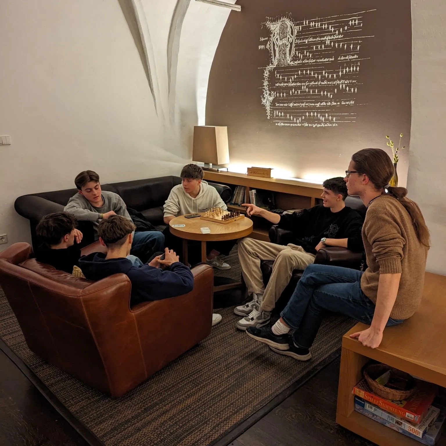 Six participants sitting around a low table with a chess set in the vaulted monastery lounge, under a wall carrying an illuminated manuscript