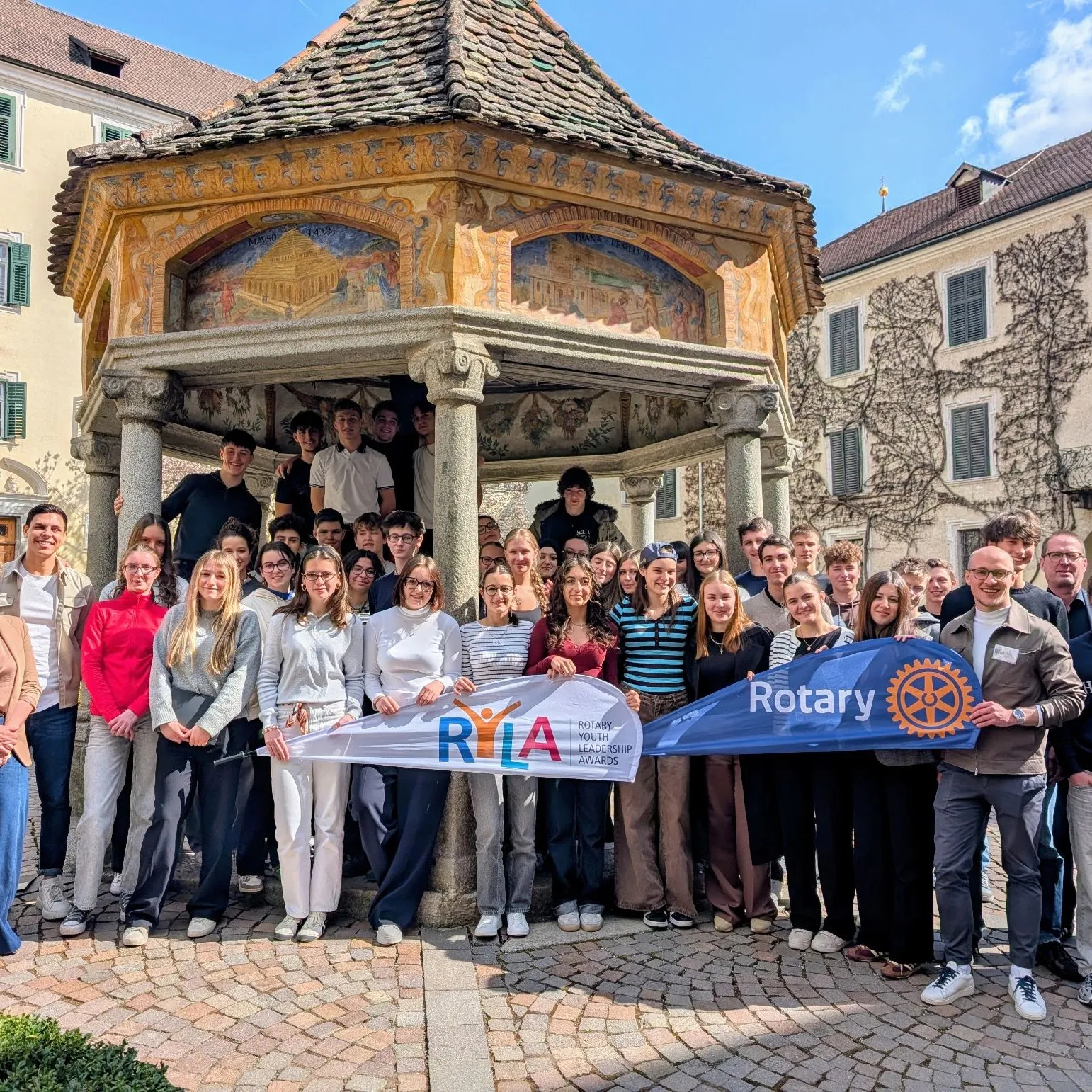 A group standing together in the monastery courtyard behind the RYLA and Rotary flags, in sunshine