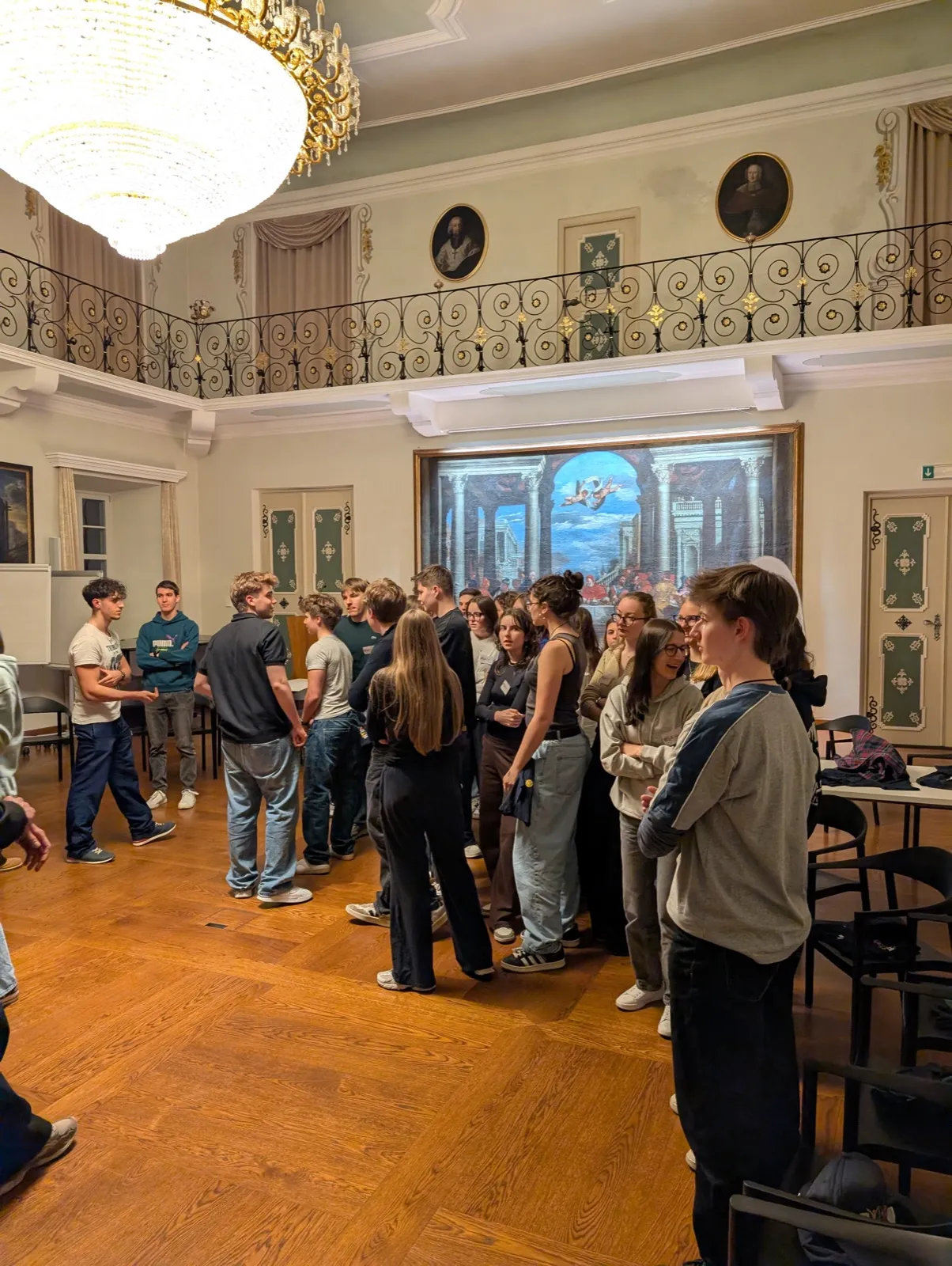 A large group of participants standing together in a hall under a chandelier, with a painting on the wall behind them