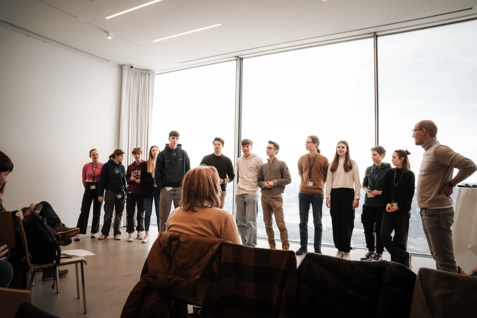 A row of participants standing in front of a floor-to-ceiling window, facing seated listeners, with an organiser standing at the end of the row