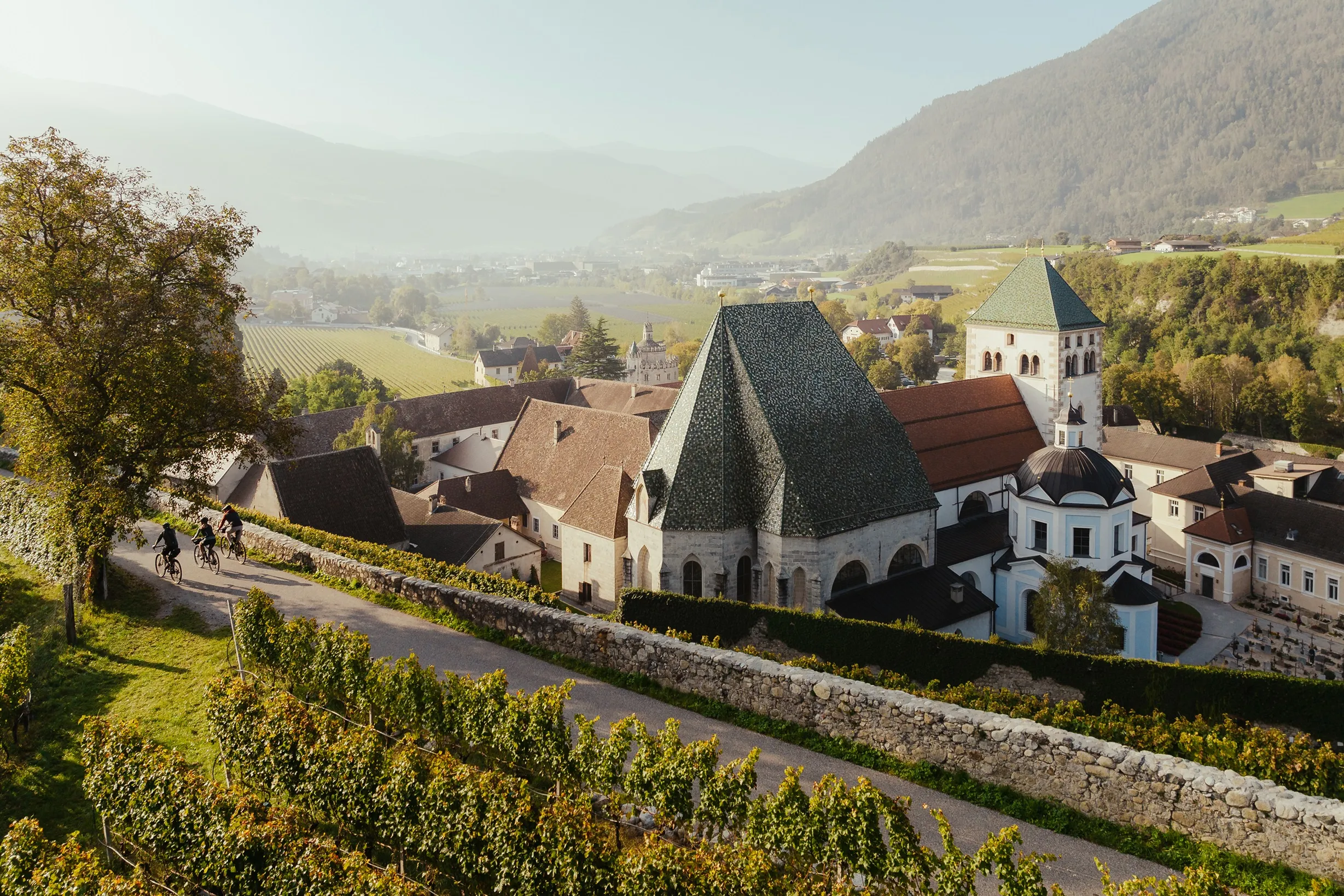 Neustift Monastery seen from the vineyard path above it: the church with its green tiled roof, the monastery buildings around it, and the valley and mountains behind