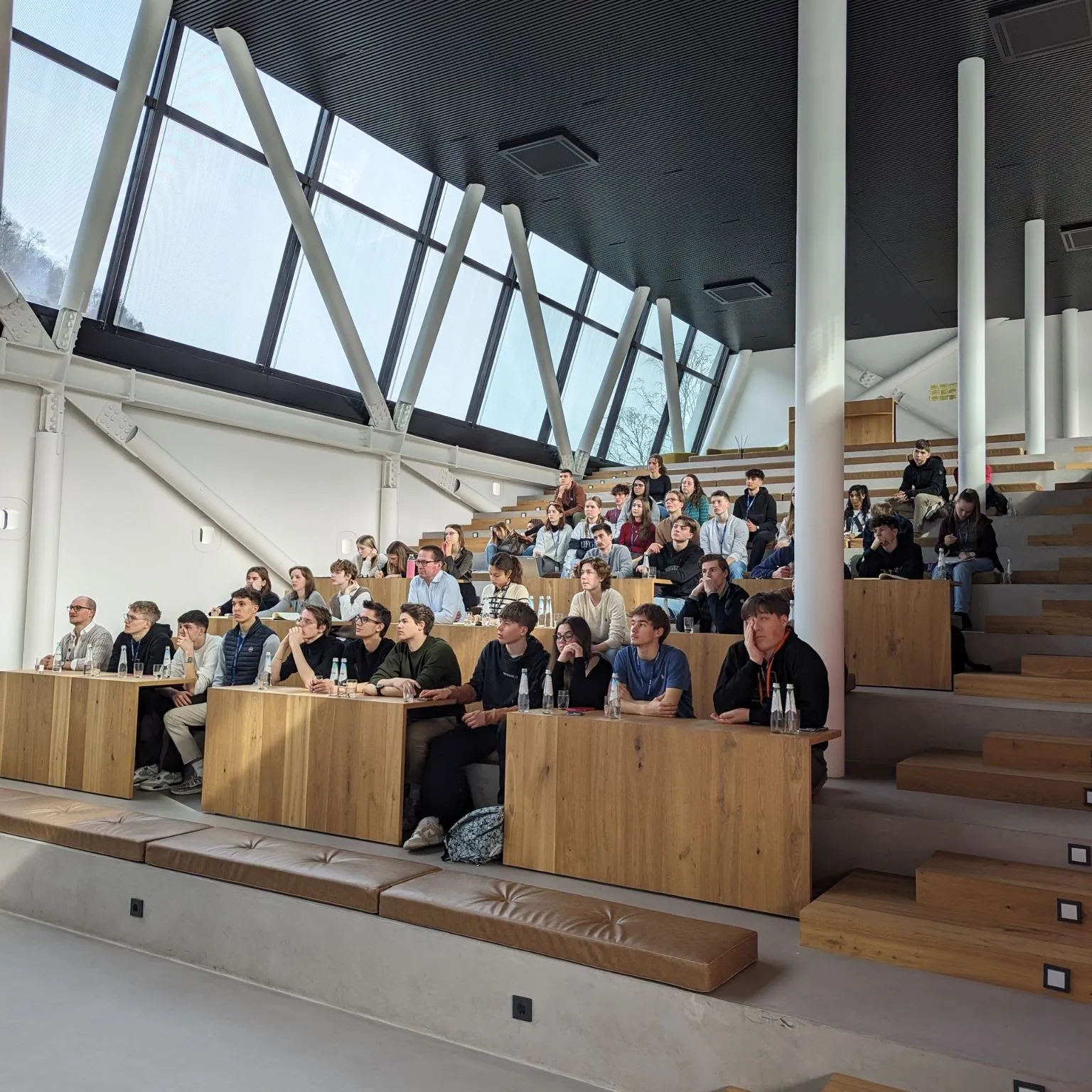 Participants seated at wooden tiered desks in a modern auditorium with a slanted glass wall, following a talk