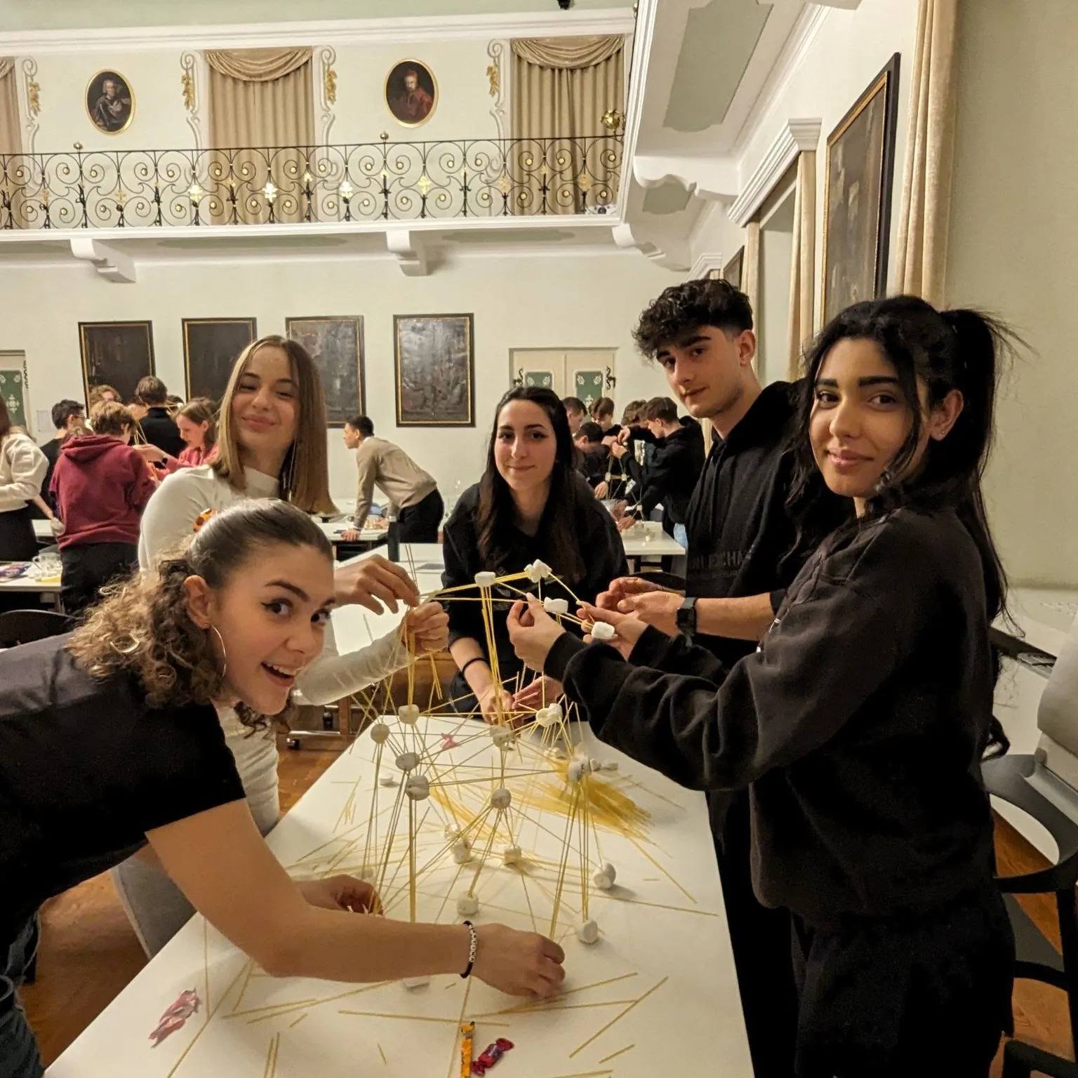 Five students in the monastery hall joining dry spaghetti with marshmallows into a spreading lattice during the Marshmallow Challenge